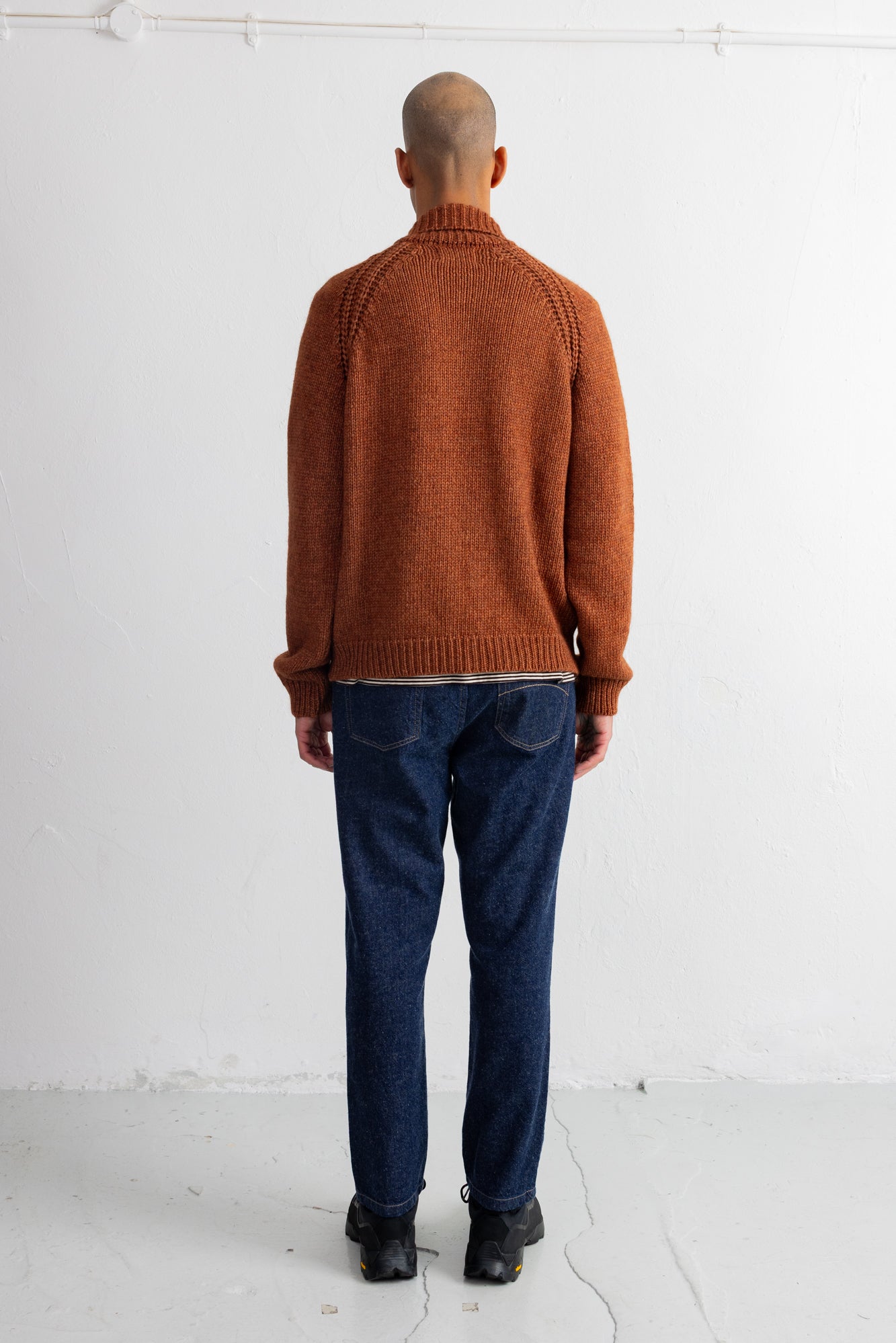 A man viewed from behind wearing a rust-orange knit sweater, dark blue jeans, and black shoes, standing against a plain white studio wall.