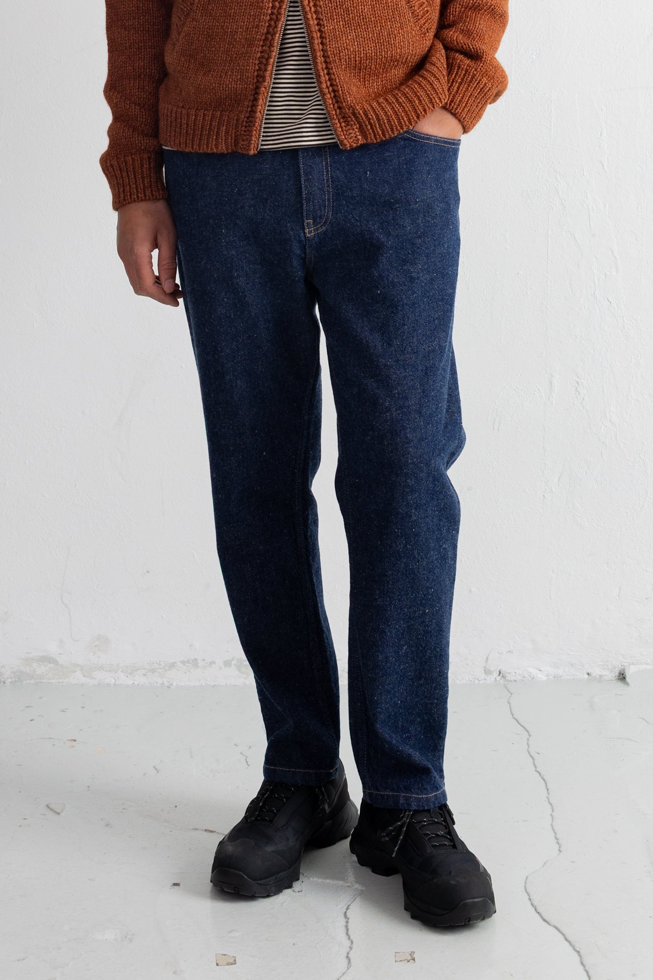 A person wearing dark blue straight-leg jeans, a rust-orange knit cardigan over a striped shirt, and black lace-up boots, standing against a white wall.
