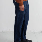 A person wearing dark blue relaxed-fit jeans, a rust-orange knit sweater, and black boots, standing against a white textured wall.