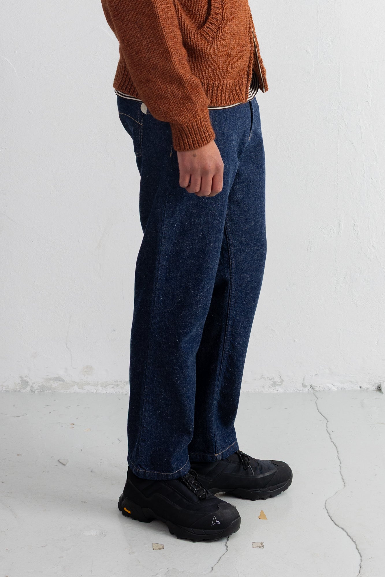 A person wearing dark blue relaxed-fit jeans, a rust-orange knit sweater, and black boots, standing against a white textured wall.