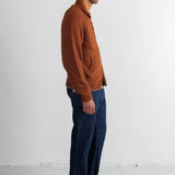 A man in profile wearing a rust-orange knit sweater, dark blue jeans, and black shoes, standing against a plain white wall.