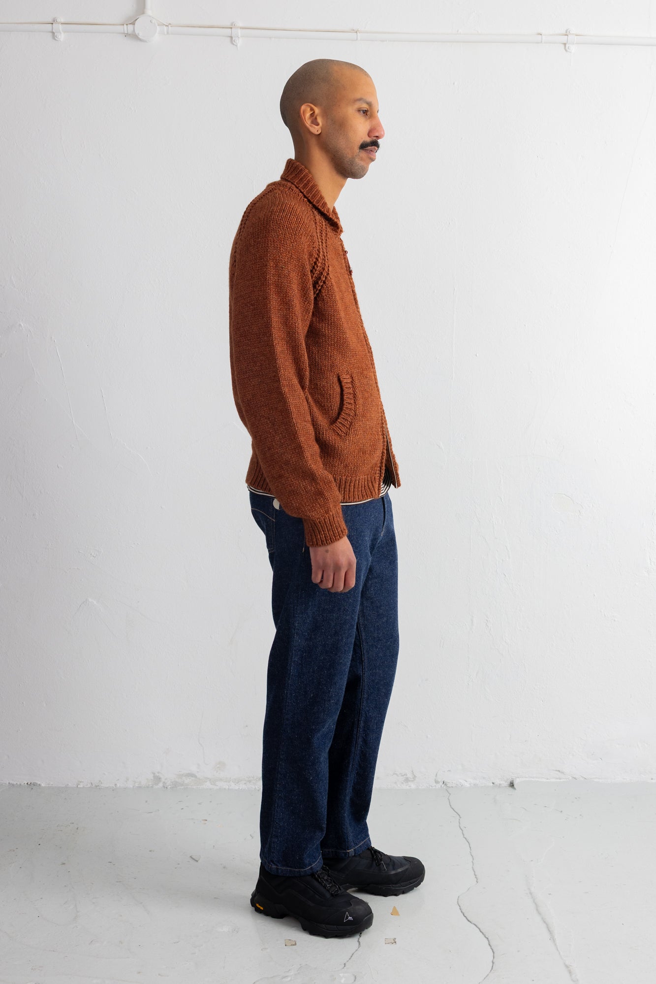 A man in profile wearing a rust-orange knit sweater, dark blue jeans, and black shoes, standing against a plain white wall.