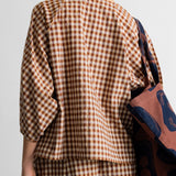 A person shown from the back wearing a brown and beige gingham check dress or outfit, with a patterned tote bag hanging over one shoulder against a plain light background.