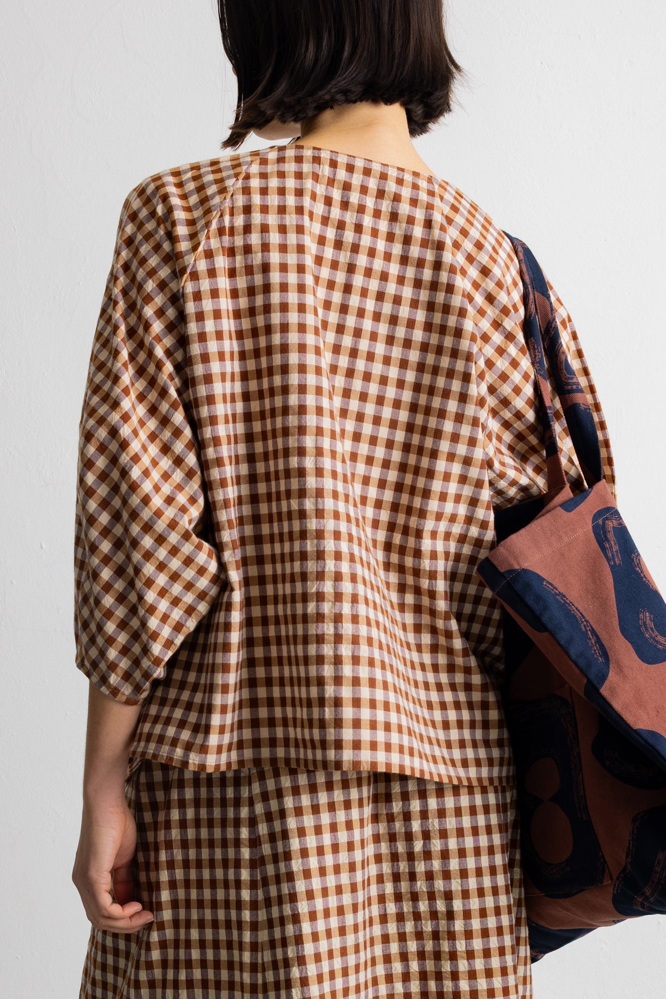A person shown from the back wearing a brown and beige gingham check dress or outfit, with a patterned tote bag hanging over one shoulder against a plain light background.
