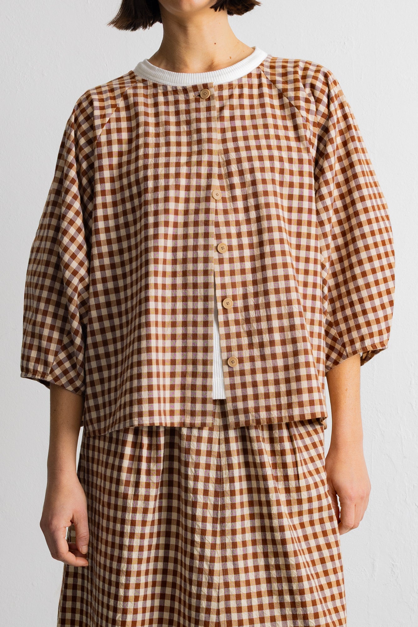 A person wearing a brown and beige gingham check dress with a white crew neck and white undershirt, shown from the neck down against a plain light background.