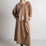 A model stands against a plain white wall wearing a brown and beige gingham check long-sleeve top and matching skirt set, styled with a white undershirt, dark socks, and beige lace-up boots.