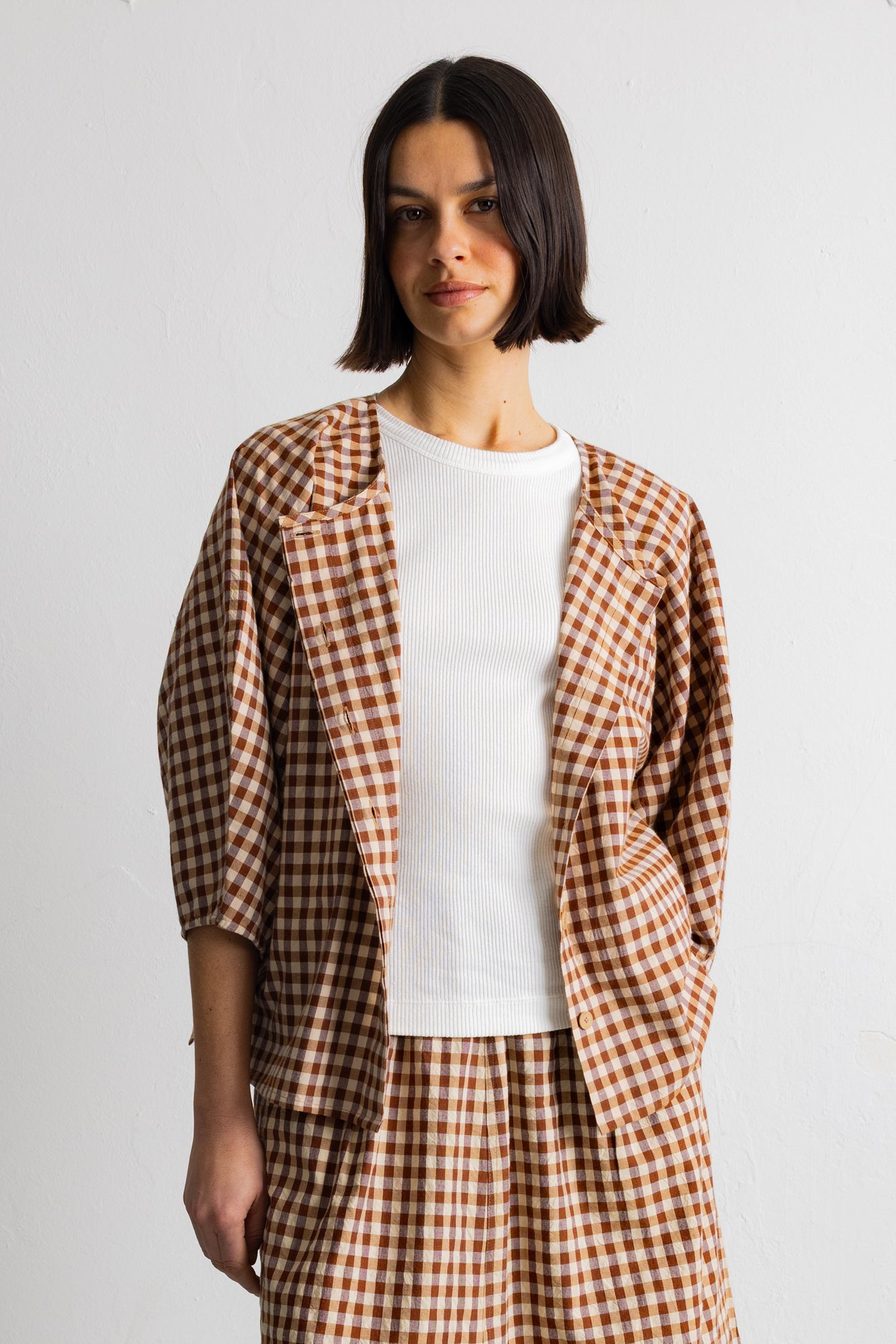 A model wearing a brown and beige gingham check blazer over a white t-shirt and matching gingham trousers, shot against a plain light gray background.