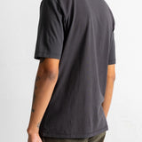 A man wearing a dark charcoal gray short-sleeve t-shirt, shown from a rear three-quarter angle against a plain white wall.
