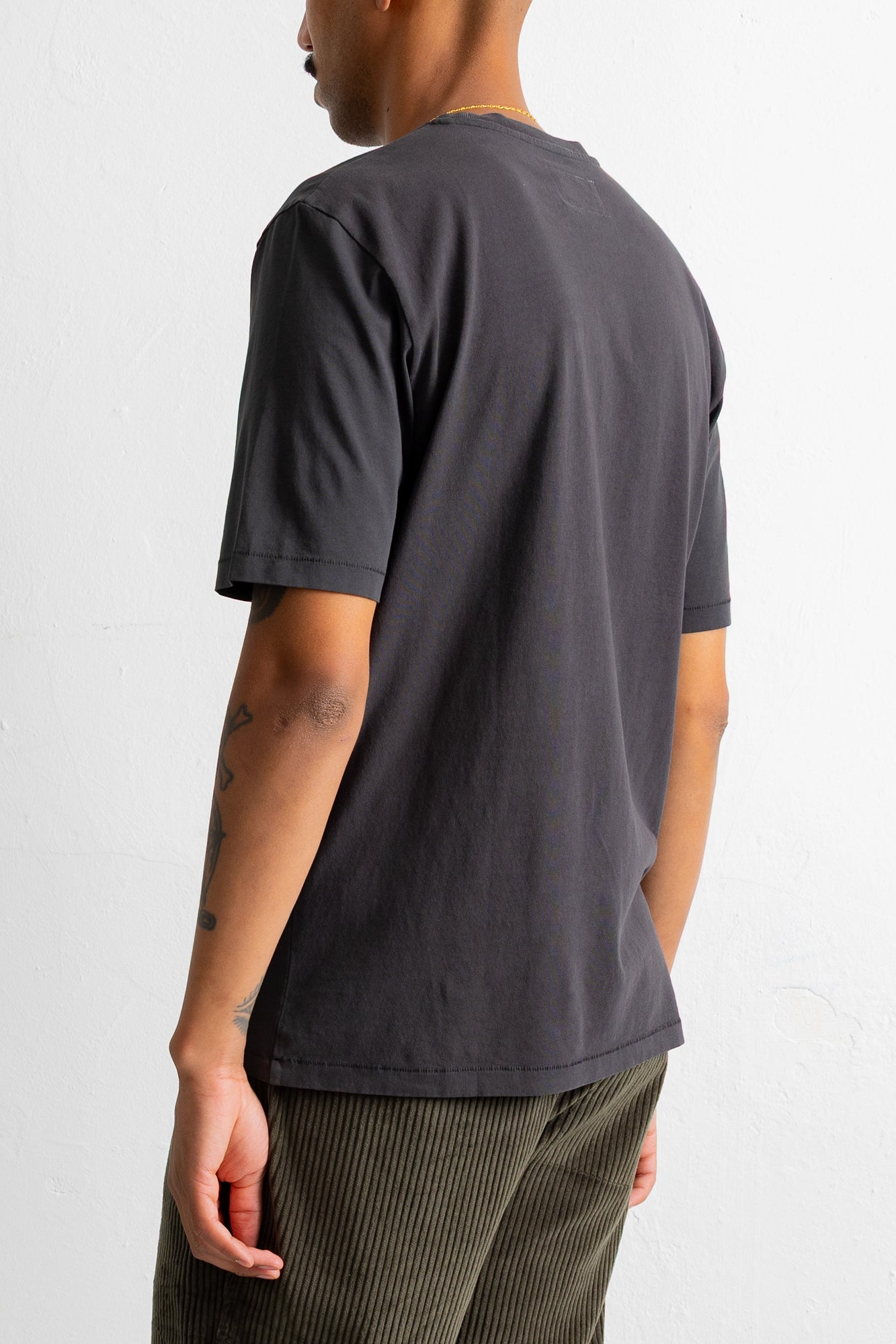 A man wearing a dark charcoal gray short-sleeve t-shirt, shown from a rear three-quarter angle against a plain white wall.