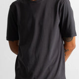 A man wearing a dark charcoal gray crew-neck t-shirt, paired with olive green corduroy pants, photographed against a plain off-white background.