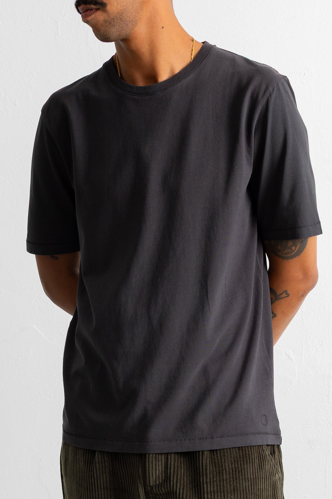 A man wearing a dark charcoal gray crew-neck t-shirt, paired with olive green corduroy pants, photographed against a plain off-white background.
