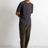 A man wearing a dark charcoal gray crewneck t-shirt, olive green corduroy pants, and black canvas sneakers with white soles, standing against a white studio wall.