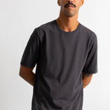 A man wearing a dark charcoal gray crew-neck t-shirt and olive green corduroy pants, standing against a plain white wall.
