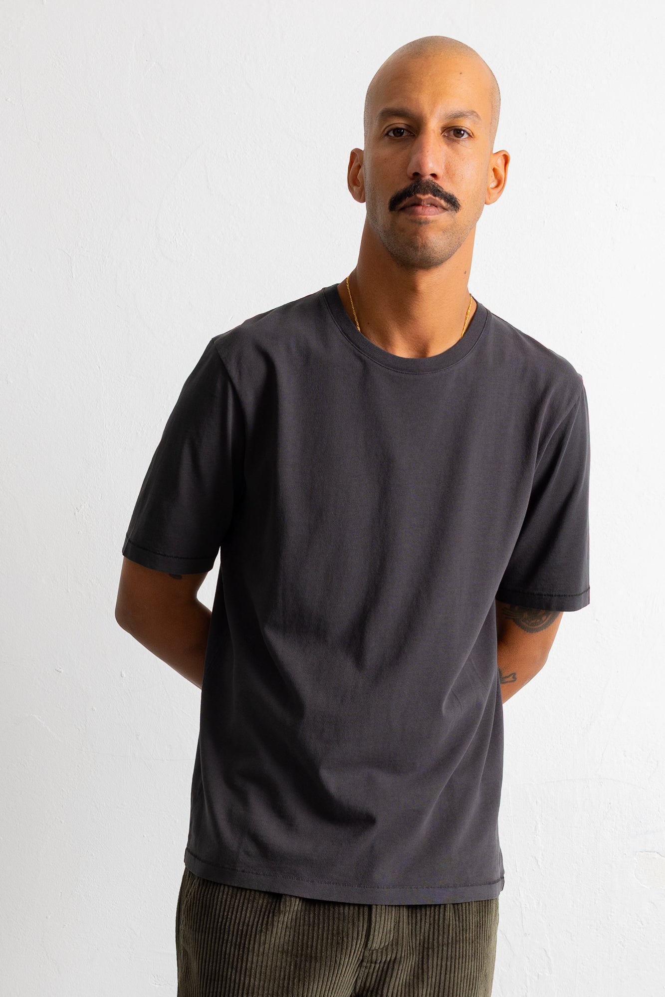 A man wearing a dark charcoal gray crew-neck t-shirt and olive green corduroy pants, standing against a plain white wall.