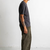 A man shown in profile wearing a dark charcoal gray t-shirt, olive green pants, and black canvas sneakers, standing against a plain white studio wall.