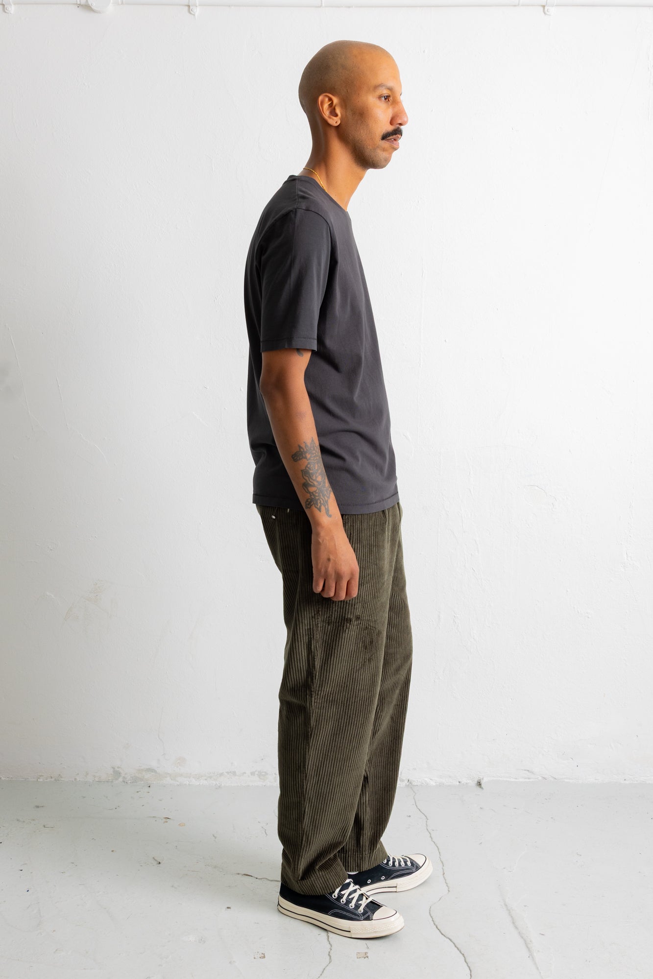 A man shown in profile wearing a dark charcoal gray t-shirt, olive green pants, and black canvas sneakers, standing against a plain white studio wall.