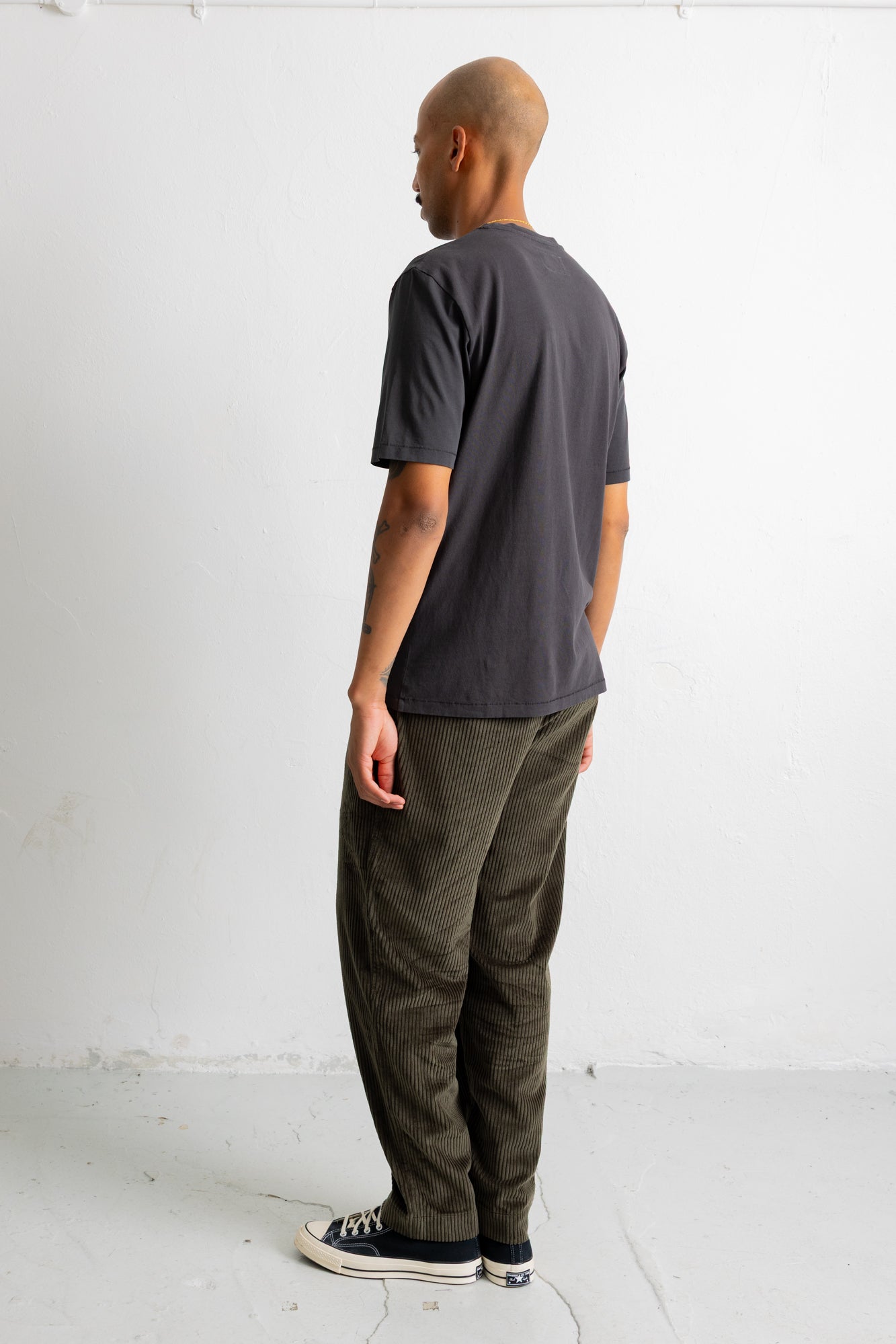 A man shown from the back wearing a dark charcoal gray t-shirt, olive green corduroy pants, and black canvas sneakers, standing against a plain white wall.