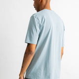 A man shown from the back wearing a light blue crewneck t-shirt and dark green pants, photographed against a plain white studio background.