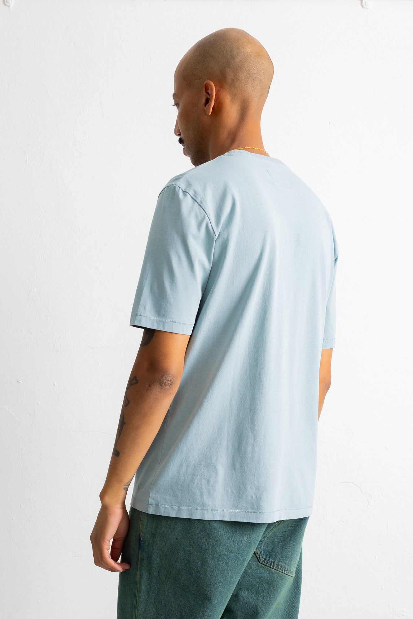 A man shown from the back wearing a light blue crewneck t-shirt and dark green pants, photographed against a plain white studio background.