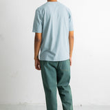 A man viewed from behind wearing a light blue crewneck t-shirt and dark green trousers, standing against a white wall.