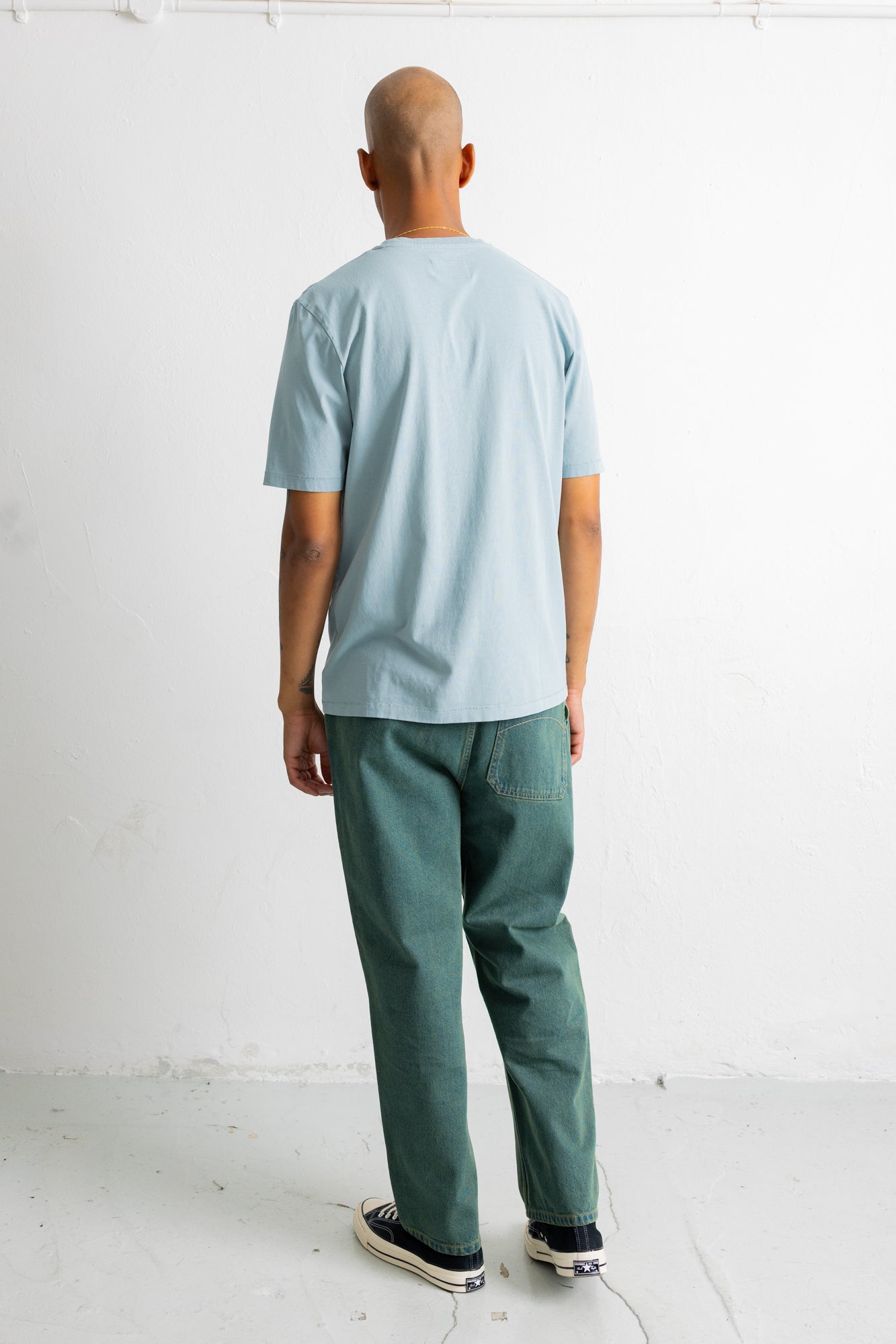 A man viewed from behind wearing a light blue crewneck t-shirt and dark green trousers, standing against a white wall.