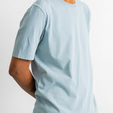 A man wearing a light blue crew-neck t-shirt and teal jeans, shown from a side profile against a plain white wall.