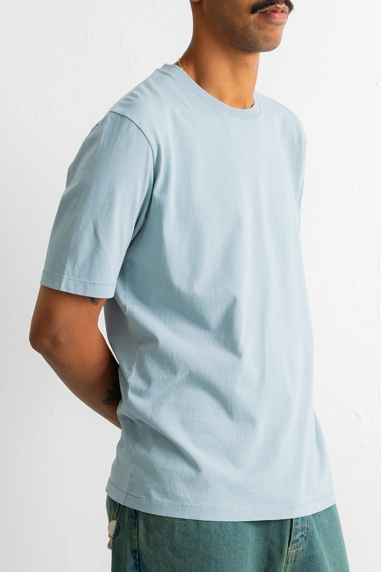 A man wearing a light blue crew-neck t-shirt and teal jeans, shown from a side profile against a plain white wall.