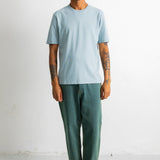 A man stands against a plain white wall wearing a light blue crew-neck t-shirt, dark green relaxed-fit pants, and black-and-white sneakers.