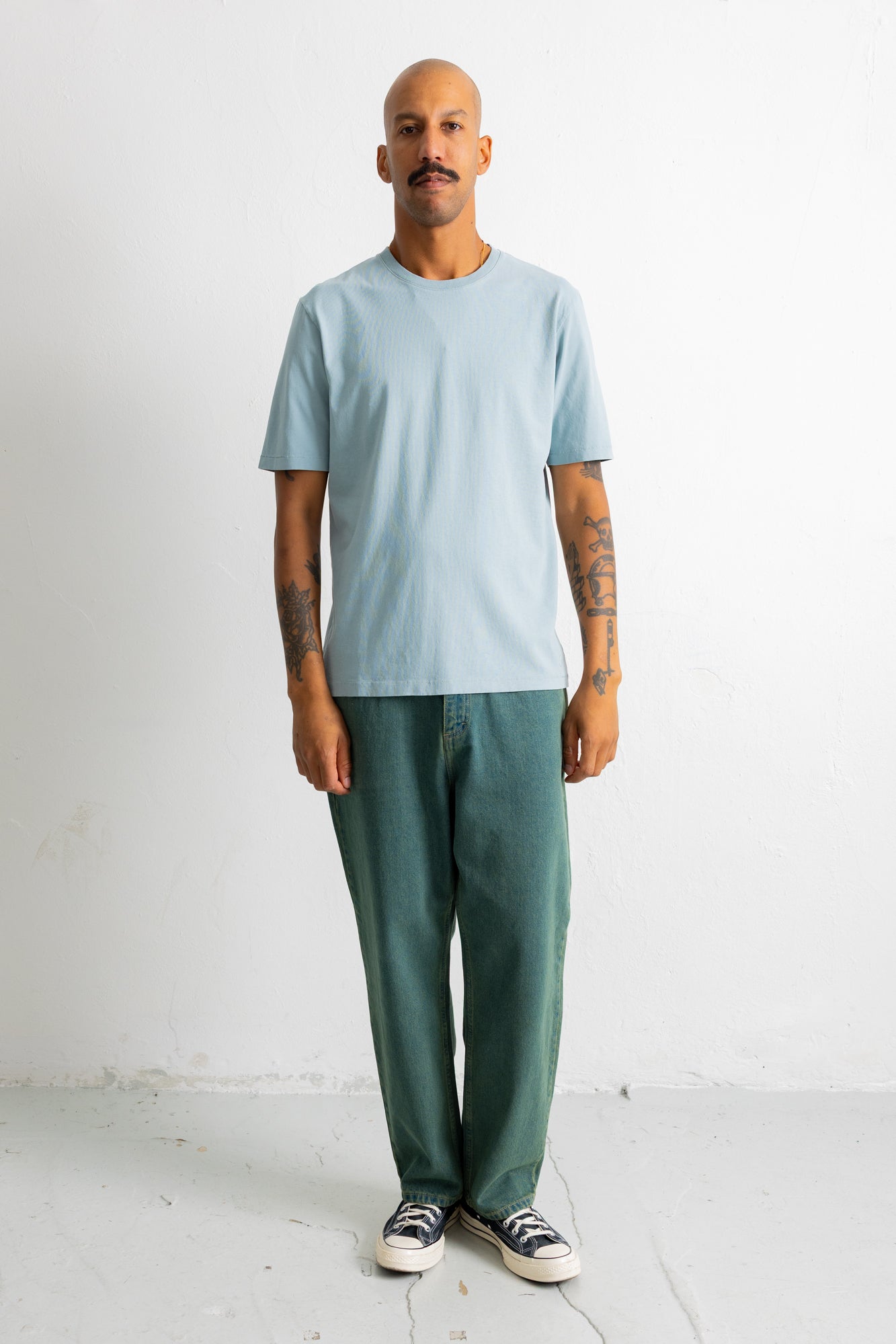 A man stands against a plain white wall wearing a light blue crew-neck t-shirt, dark green relaxed-fit pants, and black-and-white sneakers.