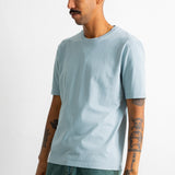 A man wearing a light blue crew-neck t-shirt and teal pants, standing against a plain white studio background.