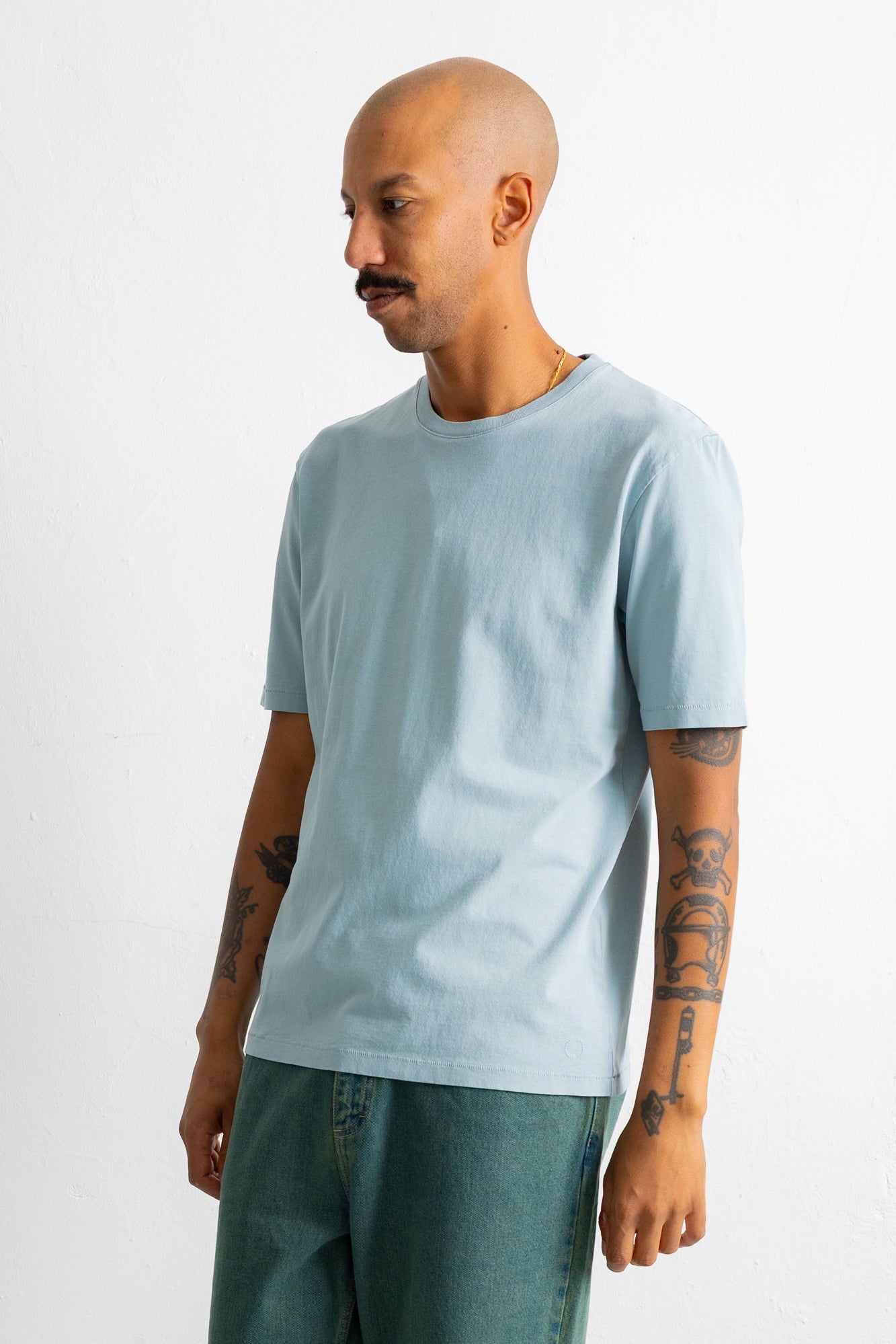 A man wearing a light blue crew-neck t-shirt and teal pants, standing against a plain white studio background.