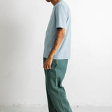 A man in profile wearing a light blue t-shirt, green wide-leg pants, and black canvas sneakers, standing against a plain white wall.