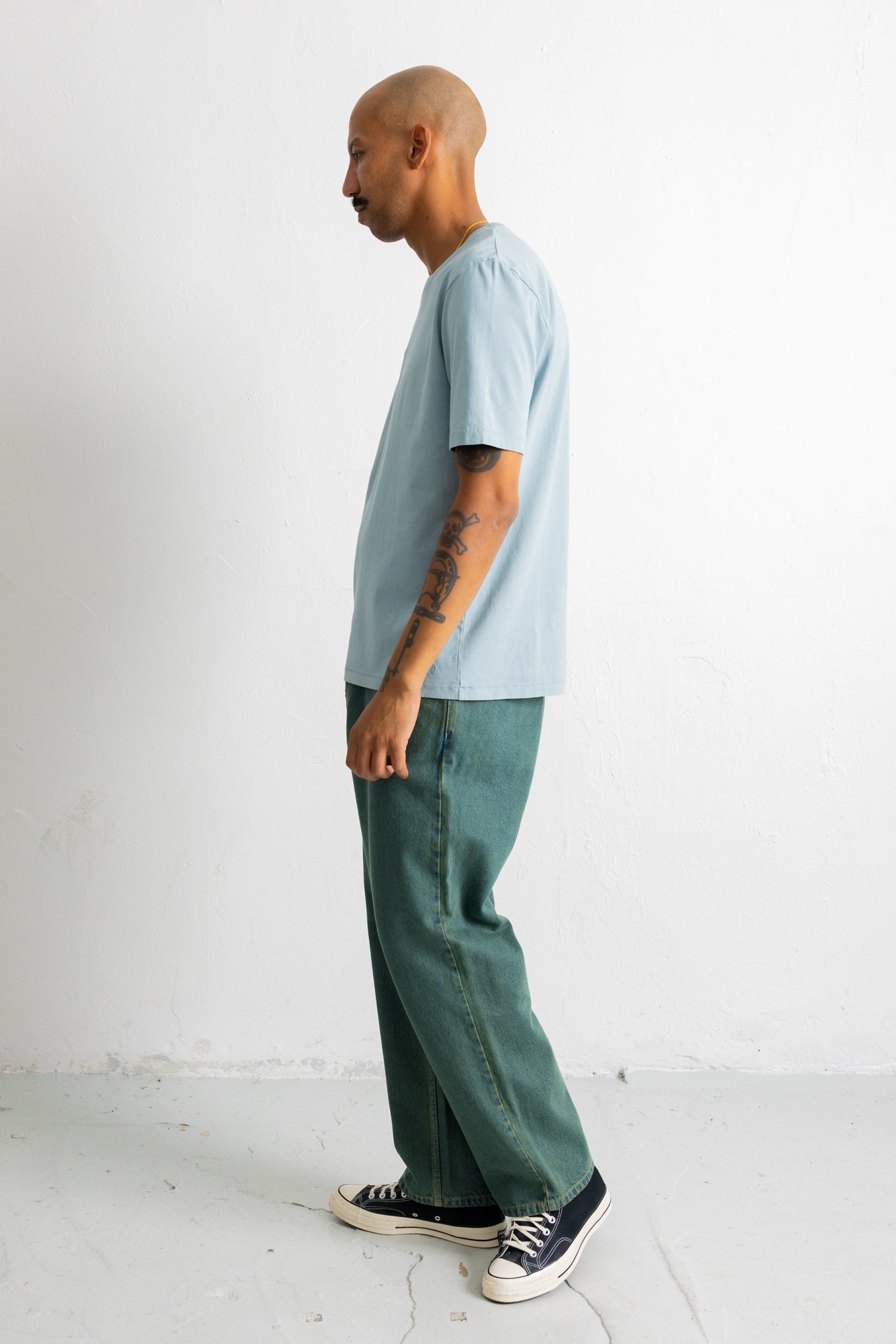 A man in profile wearing a light blue t-shirt, green wide-leg pants, and black canvas sneakers, standing against a plain white wall.