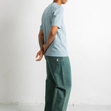 A man standing in profile wearing a light blue crewneck t-shirt, dark green wide-leg jeans, and black canvas sneakers against a plain white studio background.