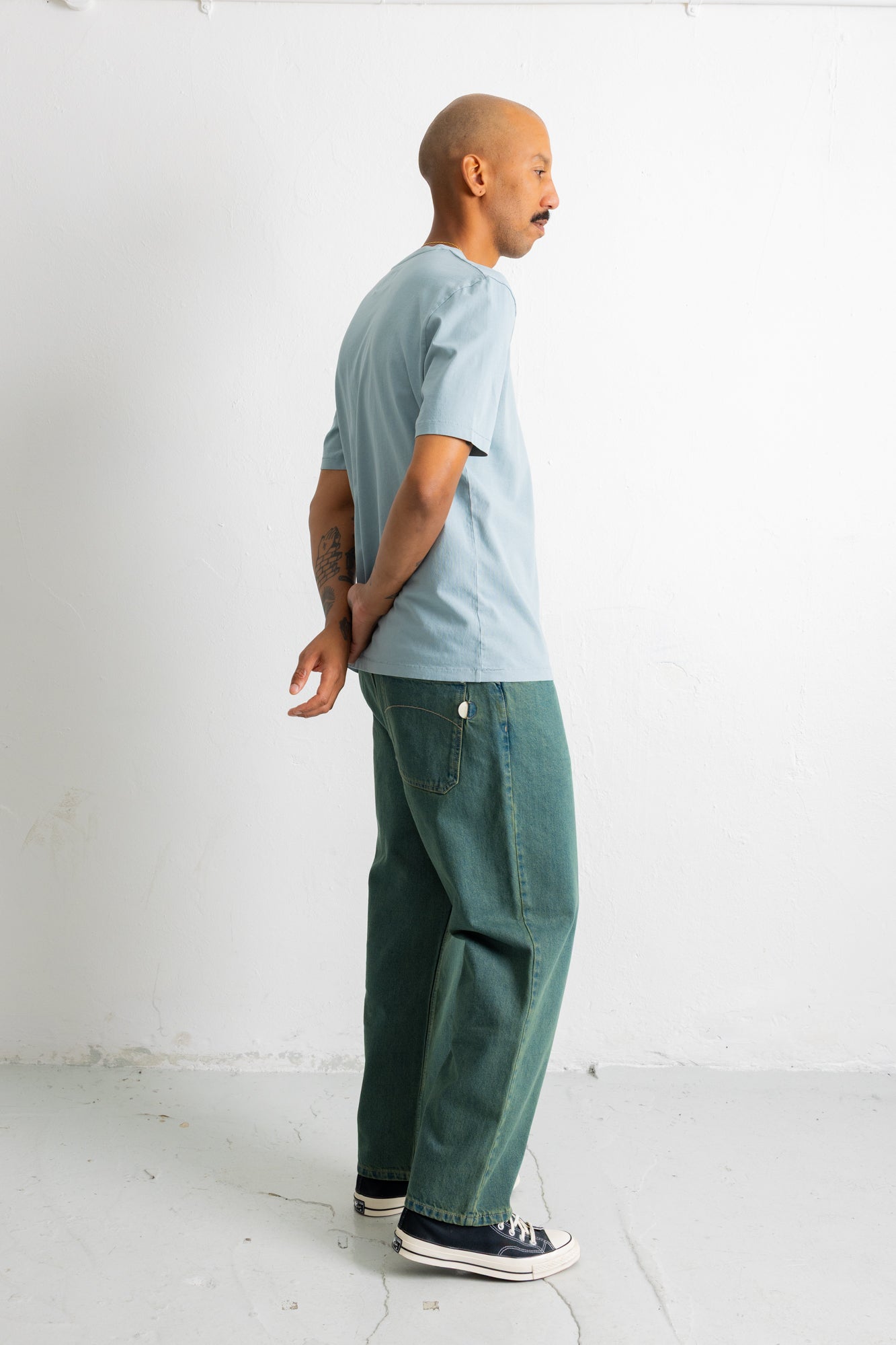 A man standing in profile wearing a light blue crewneck t-shirt, dark green wide-leg jeans, and black canvas sneakers against a plain white studio background.
