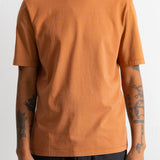 A person wearing a plain short-sleeve crew-neck t-shirt in a warm burnt orange or rust color, paired with dark pants, against a light neutral background.
