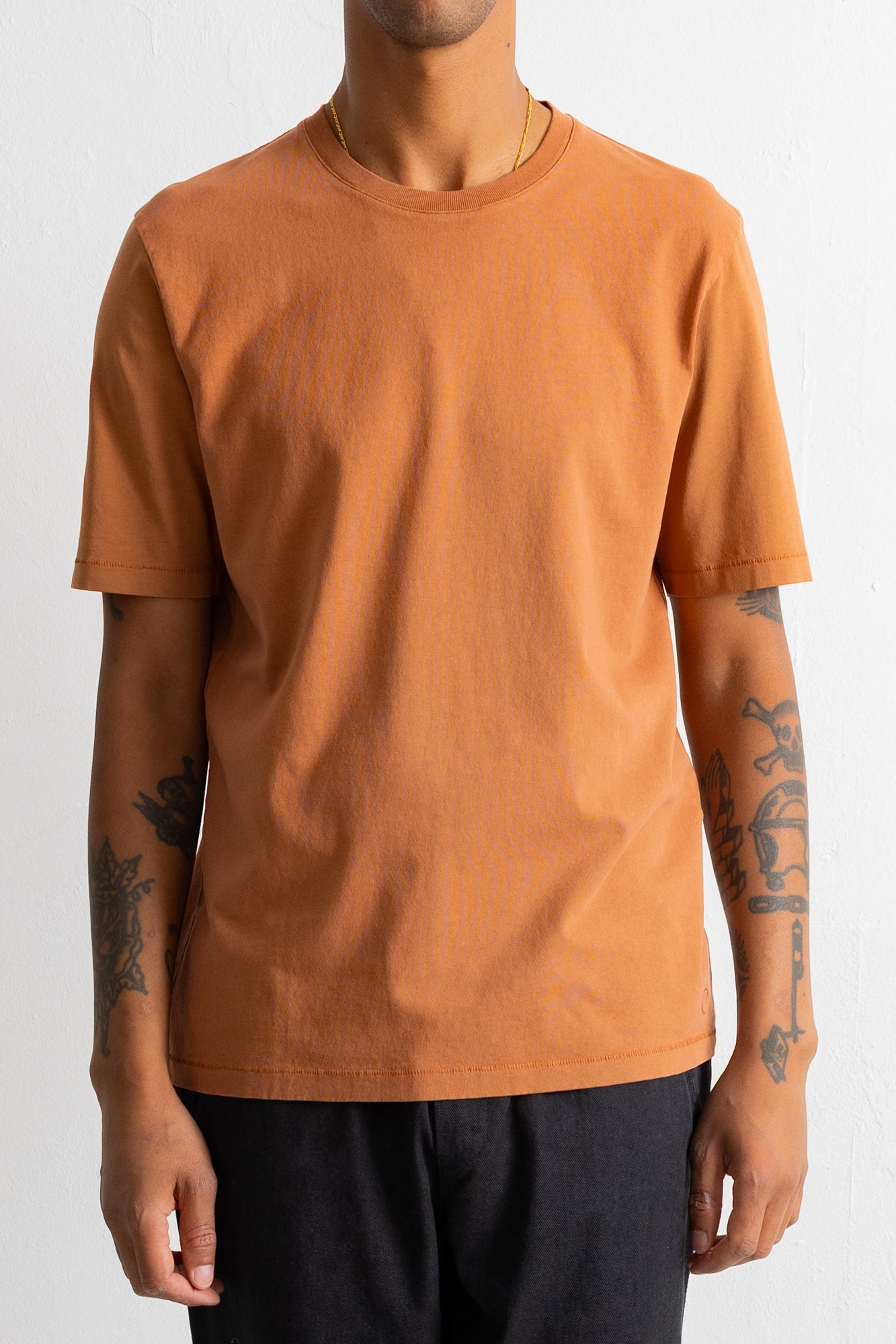 A person wearing a plain short-sleeve crew-neck t-shirt in a warm burnt orange or rust color, paired with dark pants, against a light neutral background.