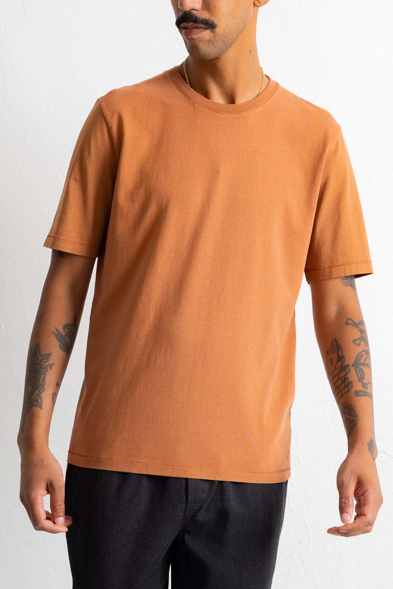 A man wearing a solid orange crew-neck t-shirt and black pants, shown from the waist up against a plain white background.
