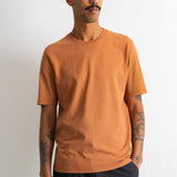 A man wearing a solid orange crew-neck t-shirt and dark pants, standing against a plain off-white wall.