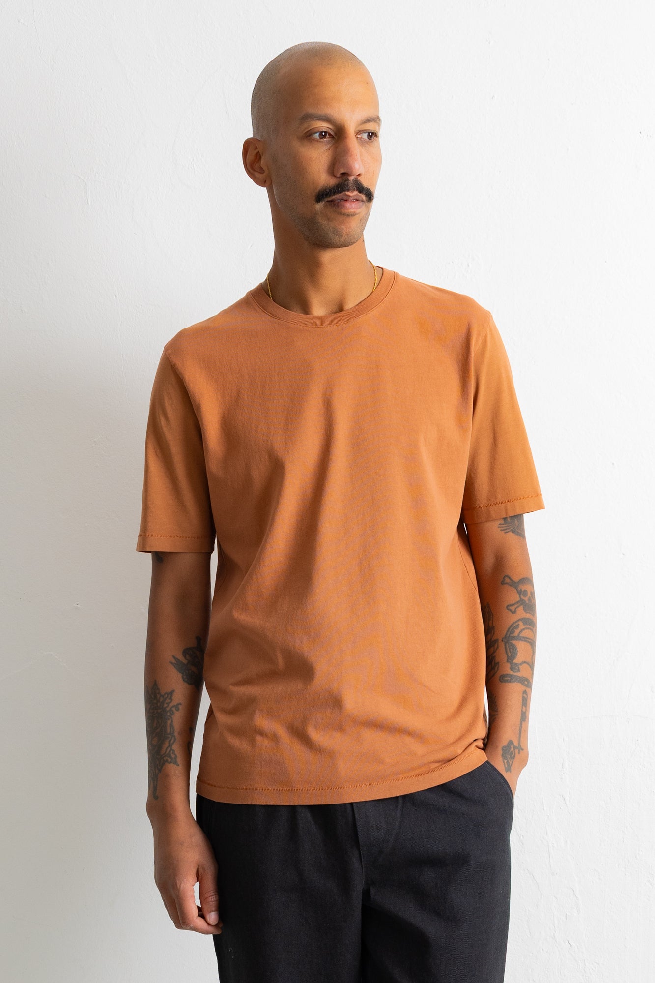 A man wearing a solid orange crew-neck t-shirt and dark pants, standing against a plain off-white wall.