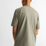 A man viewed from behind wearing a muted sage green short-sleeve t-shirt and dark blue striped pants, standing against a plain white wall.