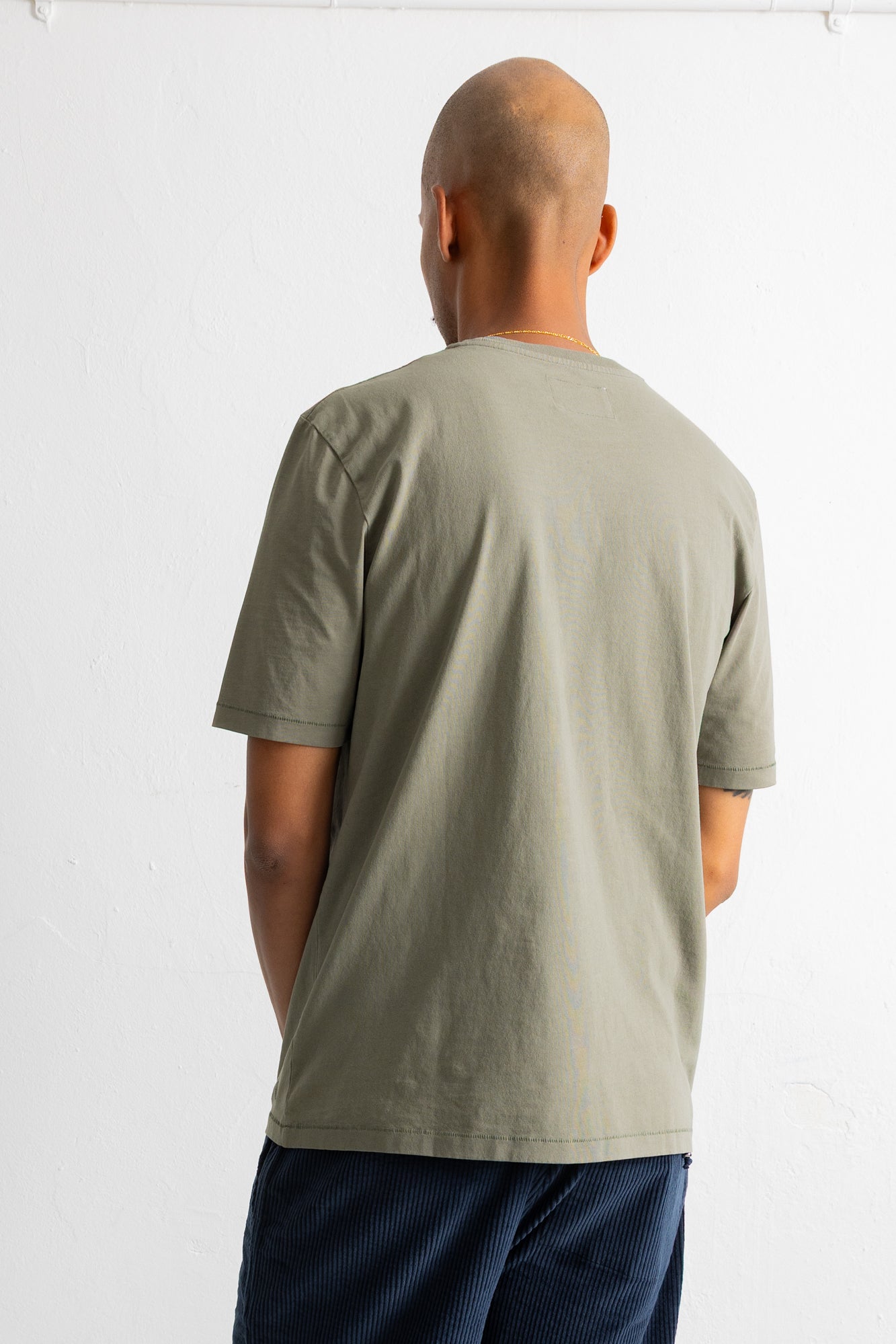 A man viewed from behind wearing a muted sage green short-sleeve t-shirt and dark blue striped pants, standing against a plain white wall.