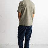 A man viewed from behind wearing a light sage green t-shirt, dark navy blue pants, and black canvas sneakers with white soles, standing against a plain white wall.
