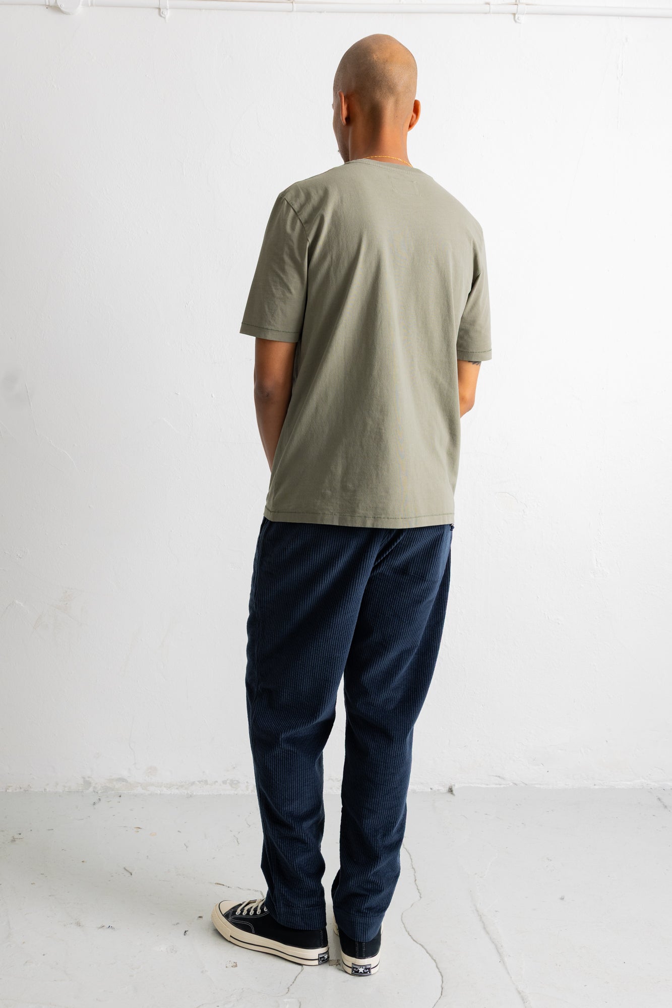 A man viewed from behind wearing a light sage green t-shirt, dark navy blue pants, and black canvas sneakers with white soles, standing against a plain white wall.
