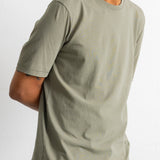 A man wearing a muted sage green crew-neck t-shirt and dark blue corduroy pants, shown from the chest down against a plain white wall.