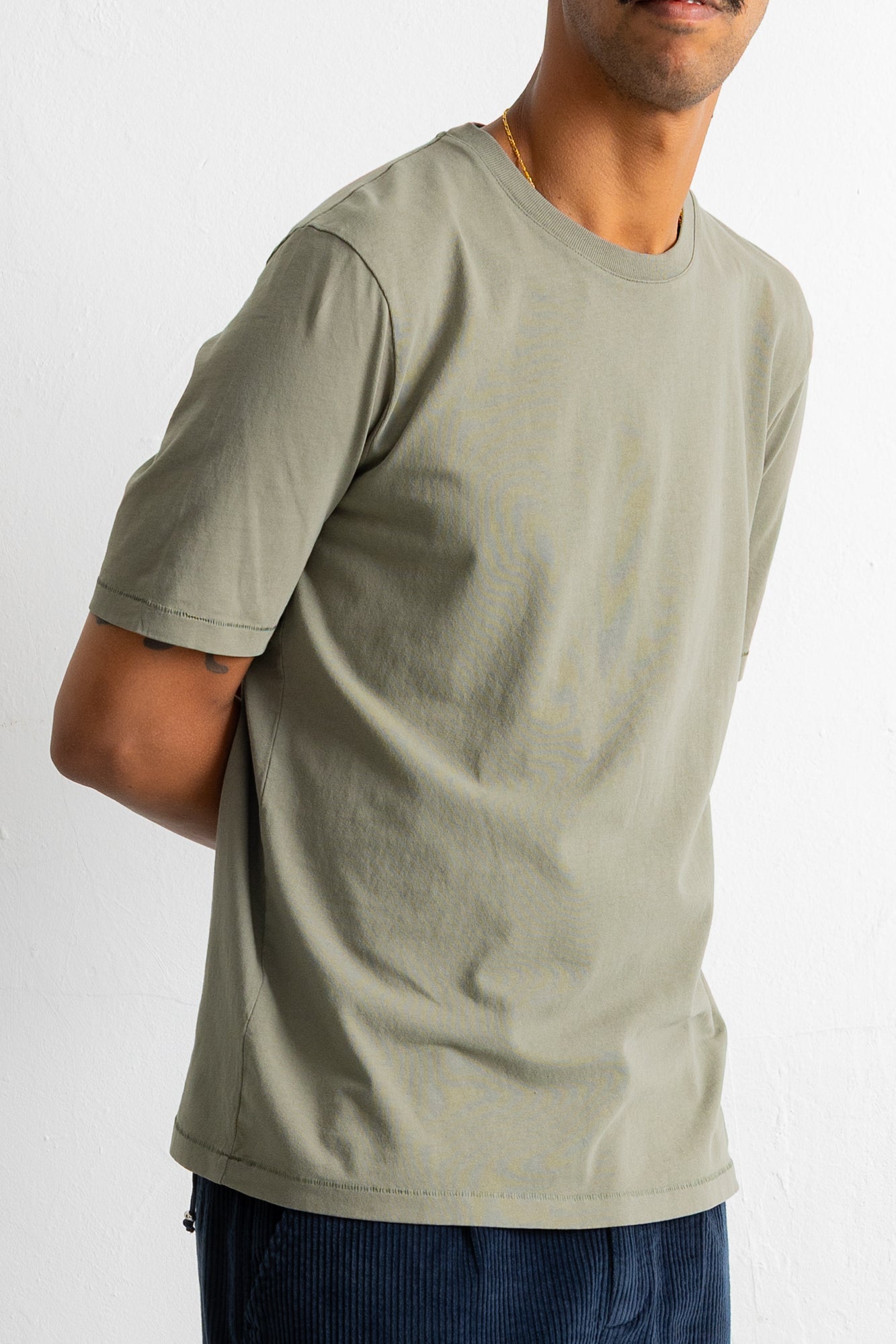 A man wearing a muted sage green crew-neck t-shirt and dark blue corduroy pants, shown from the chest down against a plain white wall.