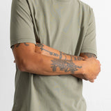 A man wearing a muted sage green crewneck t-shirt with subtle distressed paint splatters, paired with dark blue corduroy pants, shown from the neck down against a white wall.