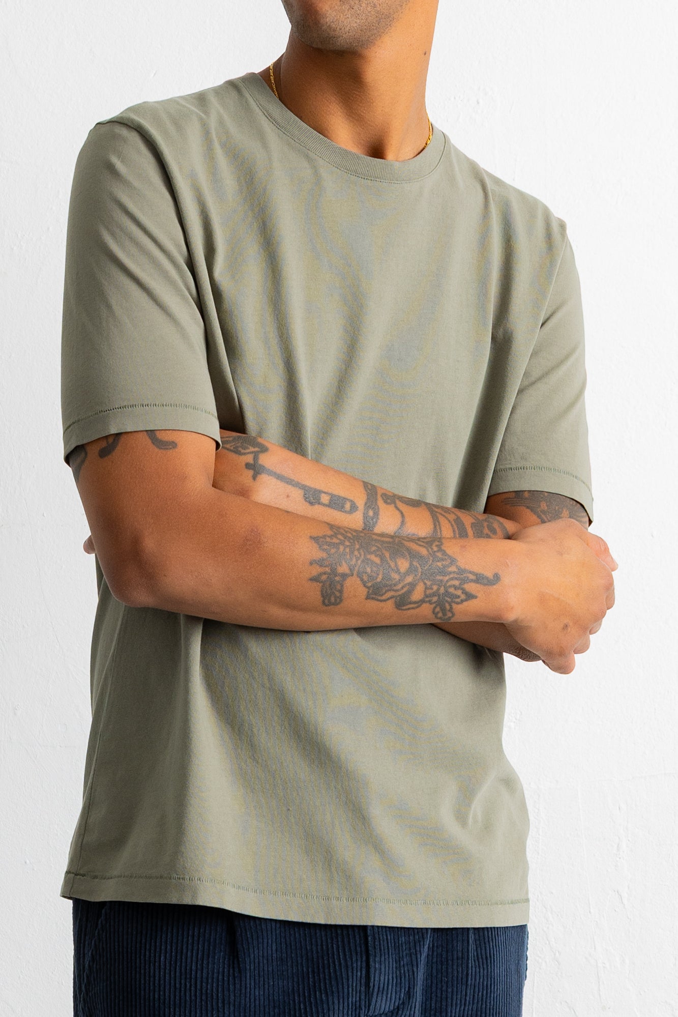A man wearing a muted sage green crewneck t-shirt with subtle distressed paint splatters, paired with dark blue corduroy pants, shown from the neck down against a white wall.