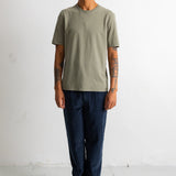 A man wearing a muted sage green short-sleeve t-shirt, dark blue jeans, and black-and-white sneakers, standing against a plain white wall.
