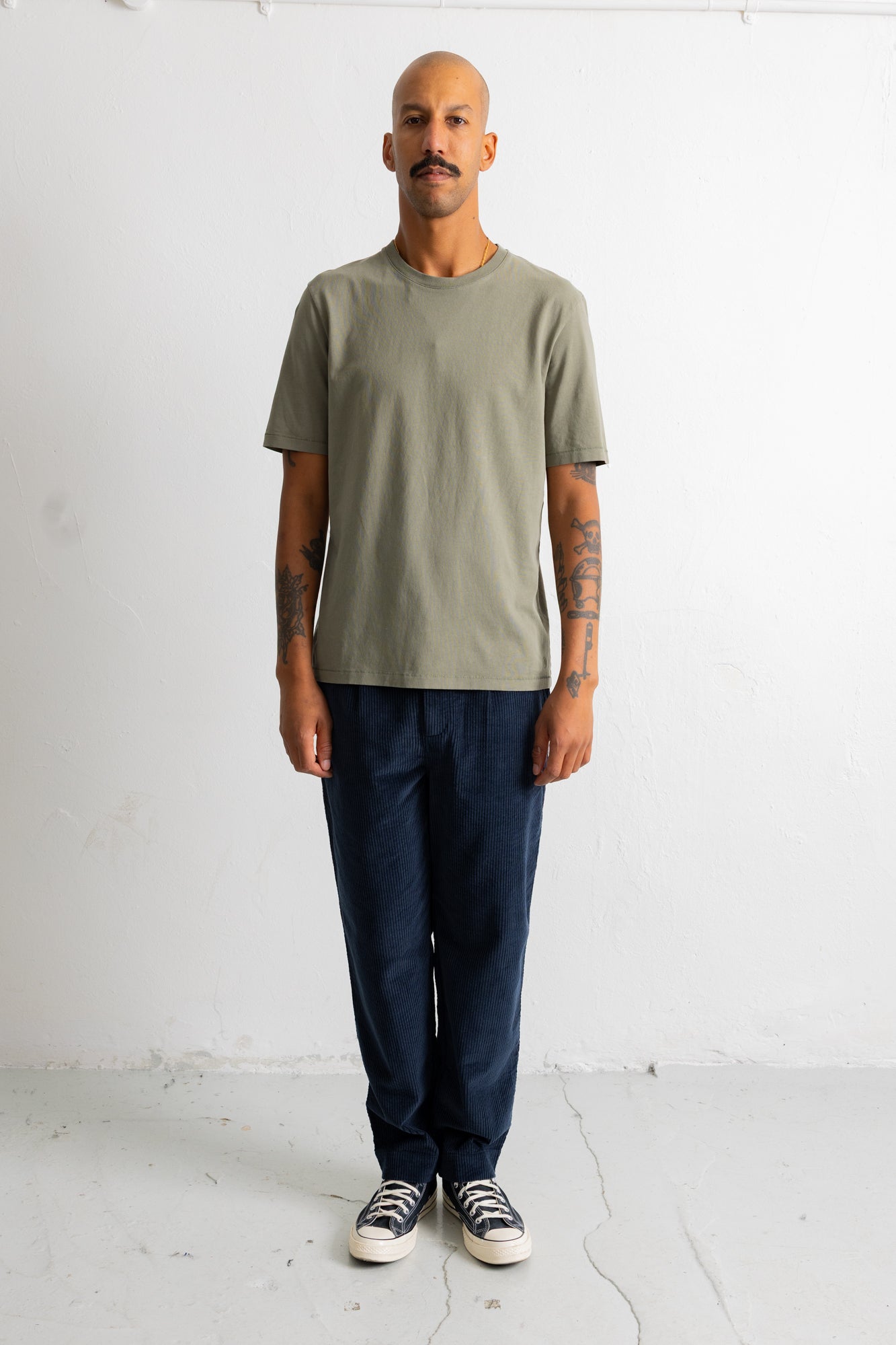 A man wearing a muted sage green short-sleeve t-shirt, dark blue jeans, and black-and-white sneakers, standing against a plain white wall.